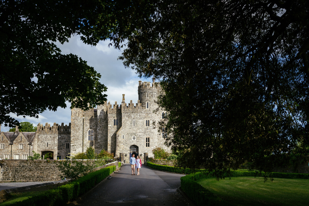 Three people walk along a path towards Kilkea Castle with crenellated towers, surrounded by greenery and shaded by trees, capturing the perfect setting for an engagement shoot.