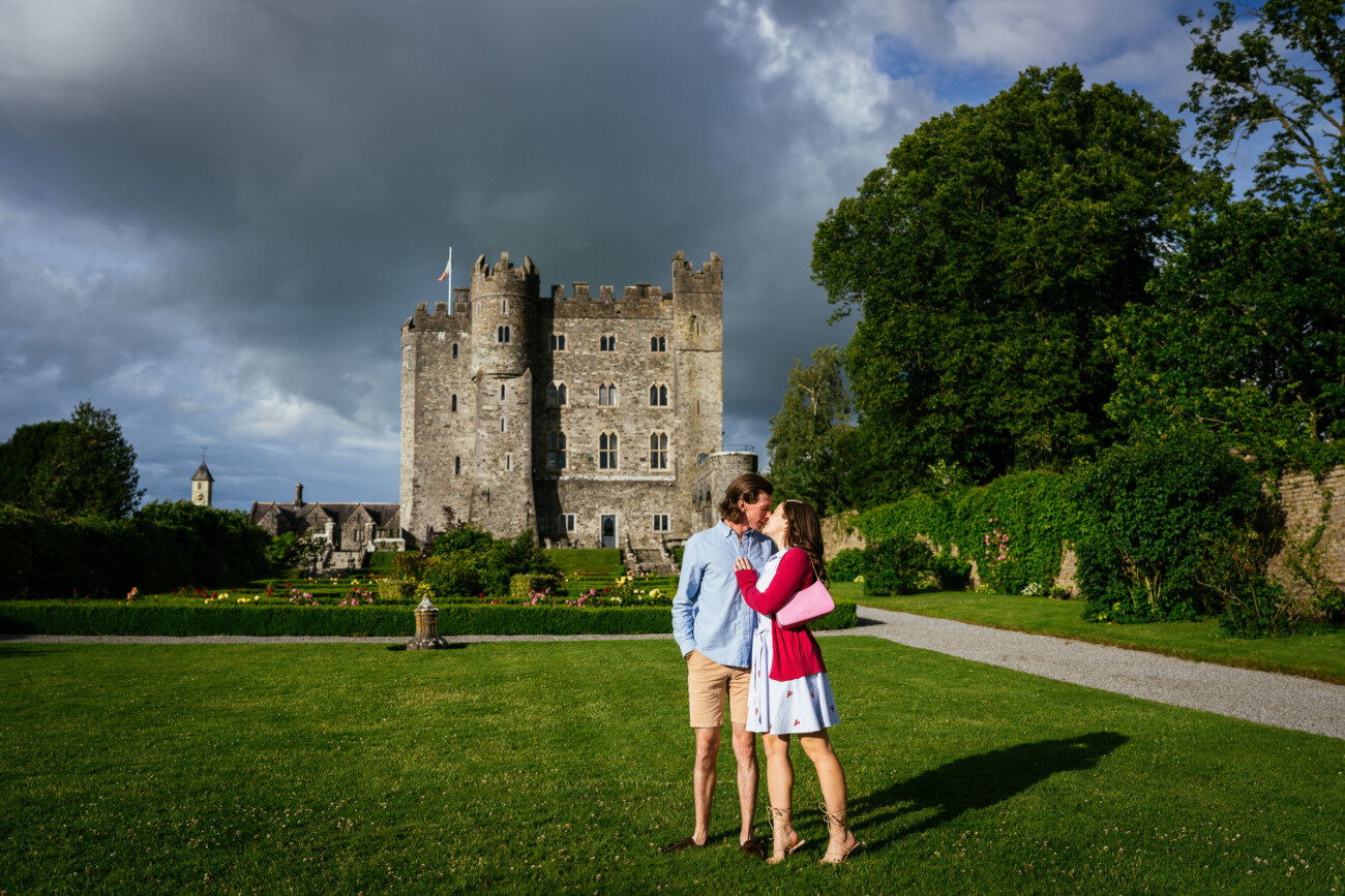 A couple stands on a lawn embracing in front of the historic Kilkea Castle under a cloudy sky, surrounded by gardens and greenery—an enchanting setting for an engagement photoshoot.