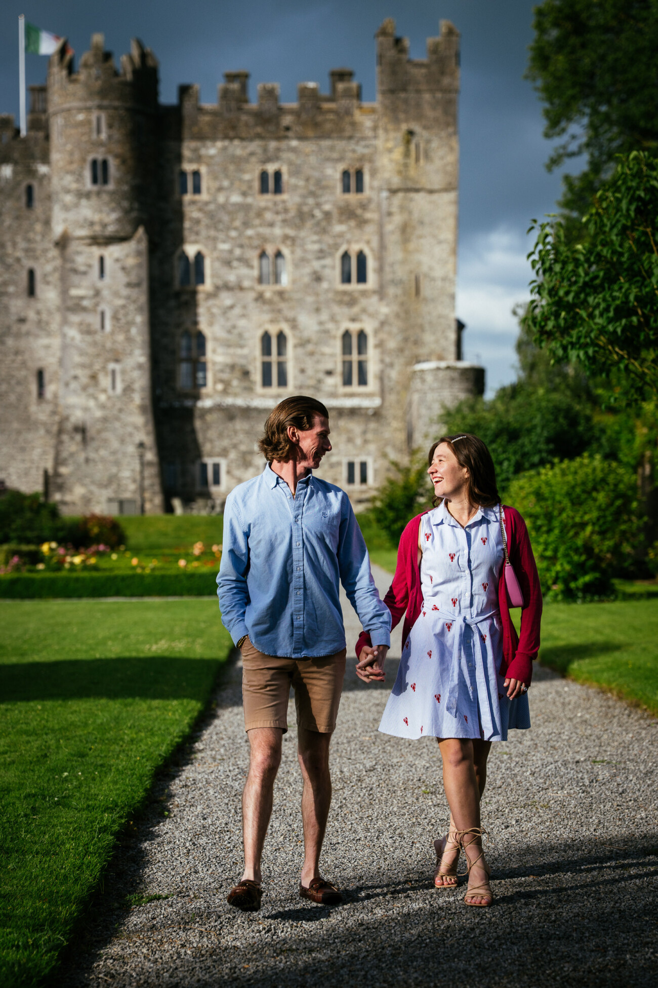 A man and woman hold hands while walking on a gravel path in front of Kilkea Castle, its stone walls framed by green lawns and gardens—the perfect setting for an engagement photoshoot.