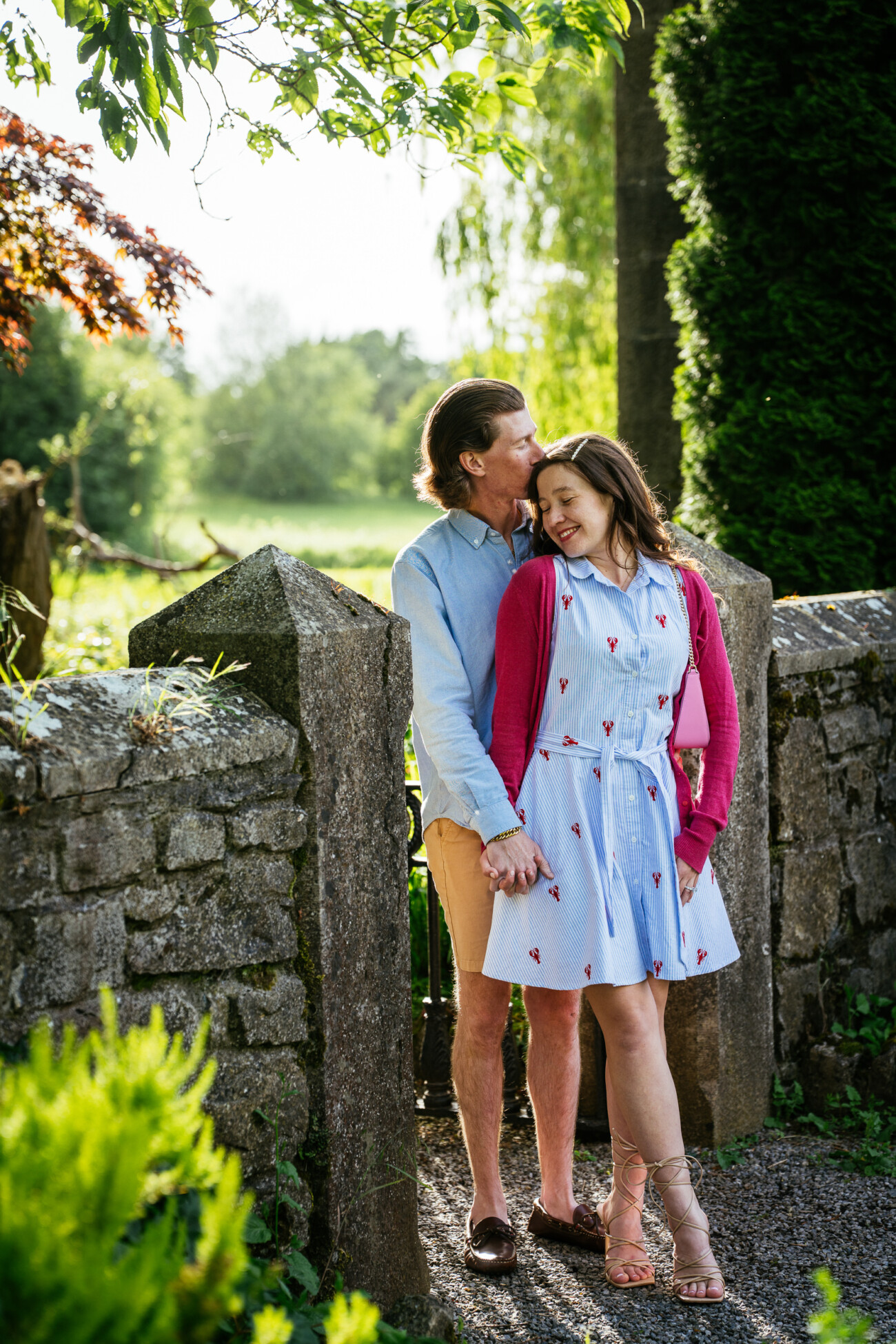 A man and woman stand in front of a stone gate at Kilkea Castle, holding hands. The man kisses the woman's head whilst she smiles with closed eyes during their romantic engagement photoshoot.