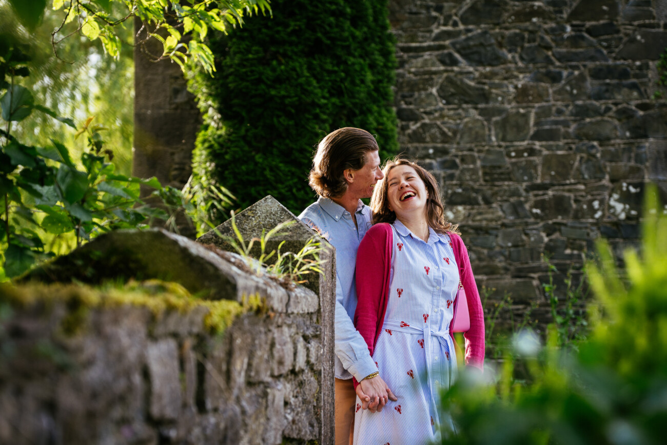 A man and woman stand close together, holding hands and smiling during their engagement photoshoot near the old stone wall and greenery of Kilkea Castle.