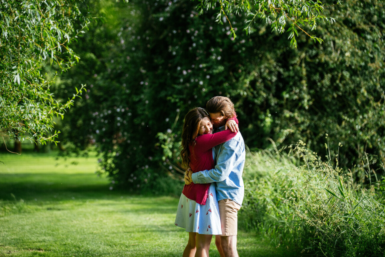 A man and woman hug each other in a grassy, sunlit park surrounded by green trees and bushes, capturing a sweet moment during their engagement photoshoot.