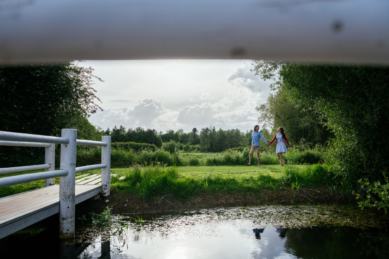 Two people holding hands walk on a grassy field near a pond at Kilkea Castle, partially framed by a white wooden bridge in the foreground—an idyllic setting for an engagement photoshoot.