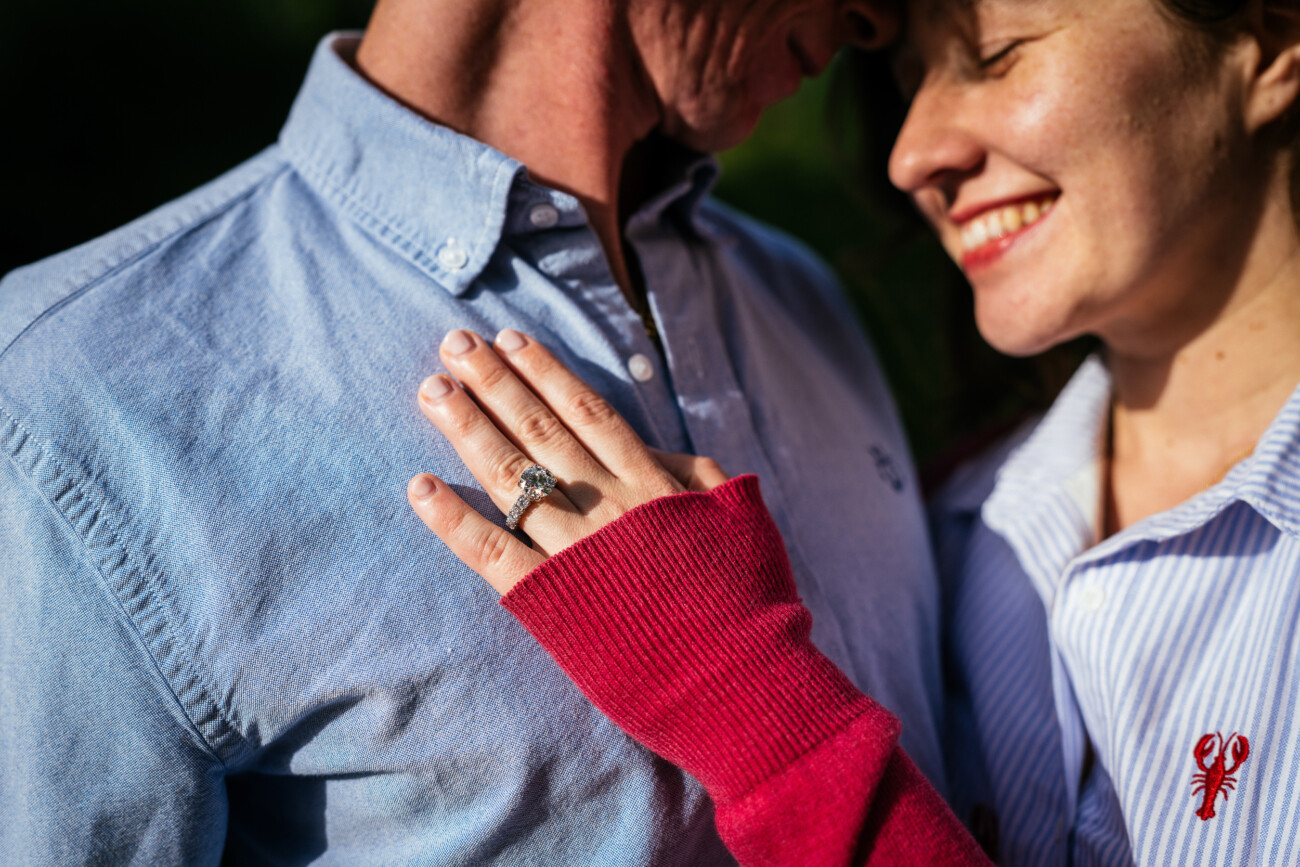 A smiling couple lean close together during their engagement shoot. The person on the right shows an engagement ring on their hand, resting on the other's chest. Both wear blue shirts; one has a red jumper sleeve.