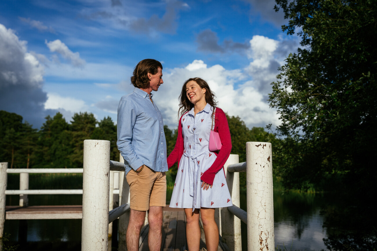 A man and a woman stand together on a wooden bridge outdoors, smiling at each other during their engagement photo shoot, with trees and a blue sky in the background.
