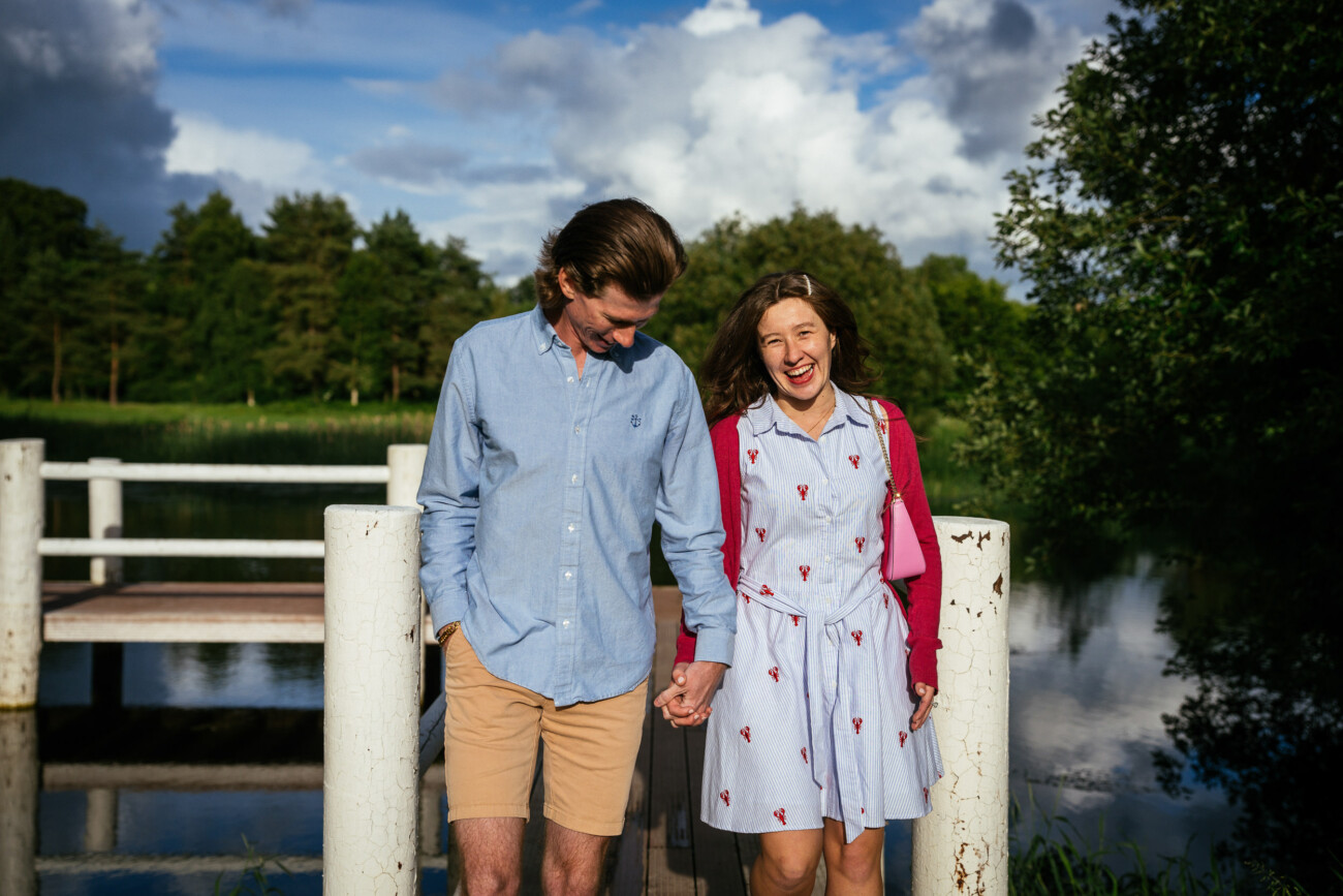 A man and woman hold hands and walk along a wooden jetty near water, surrounded by trees and greenery, during their sunny engagement photoshoot.