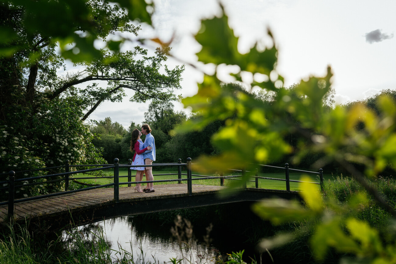 Two people stand close together on a small wooden bridge over a stream, surrounded by green trees and foliage, with leaves partially framing the foreground—capturing the intimate moment of their engagement proposal.