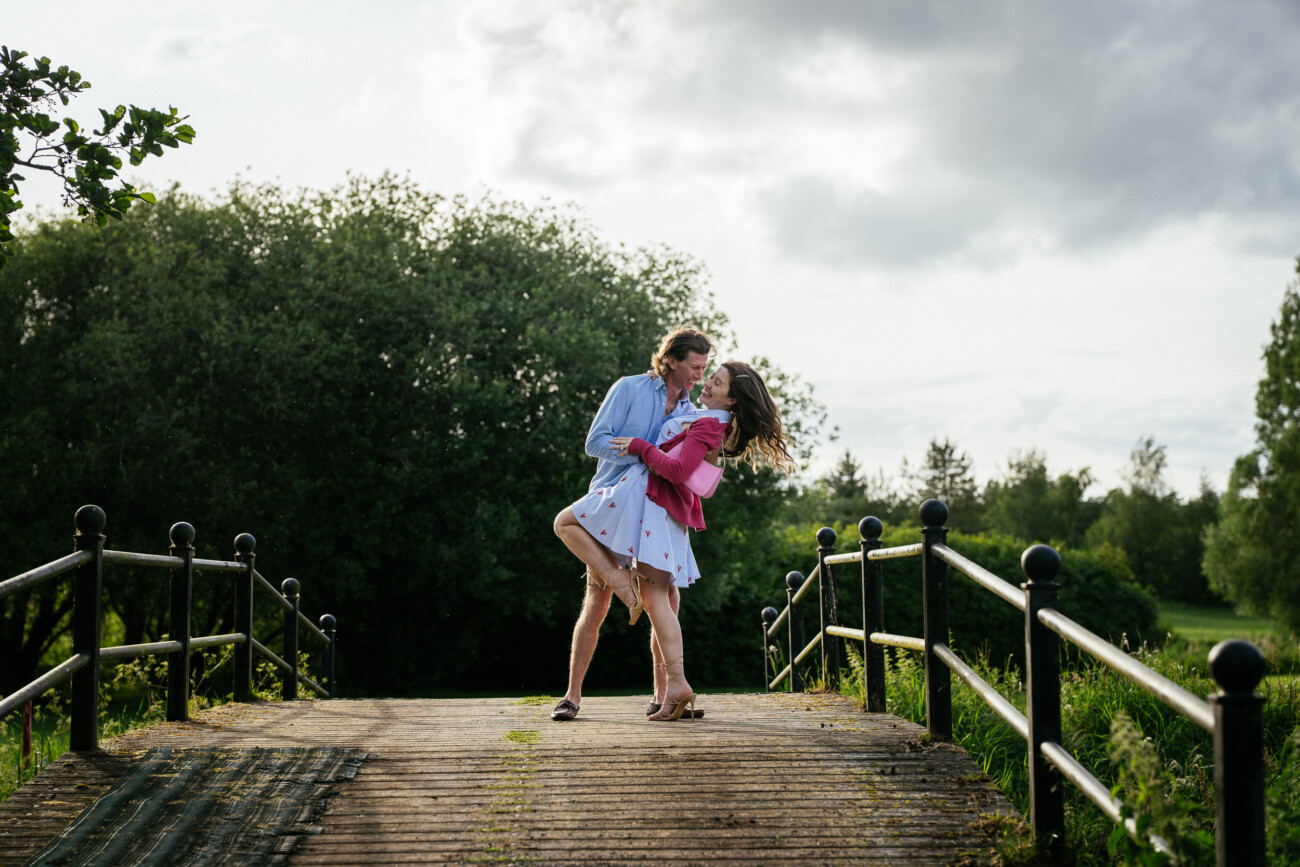 A couple embraces and kisses on a small wooden bridge surrounded by greenery under a partly cloudy sky, celebrating their engagement.
