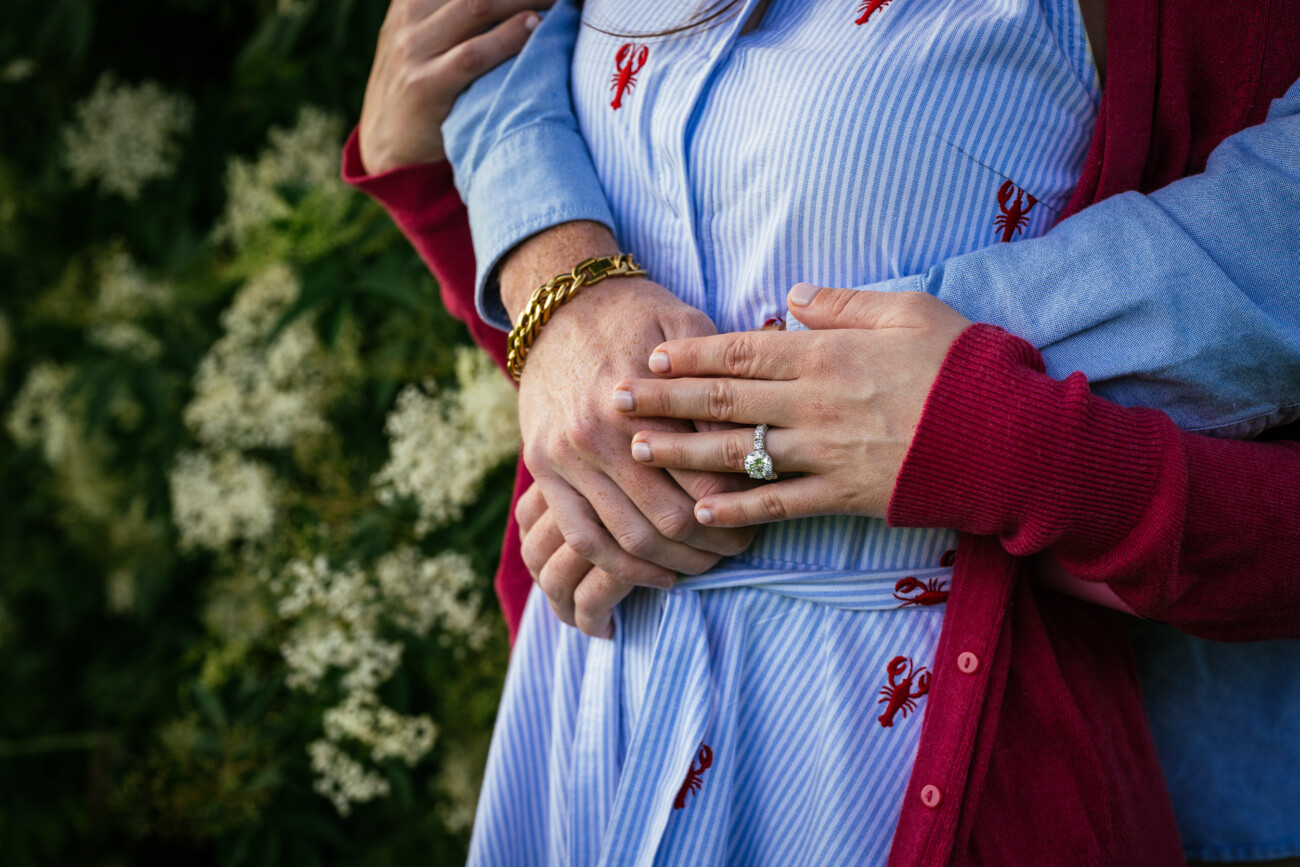 A person wearing a red jumper shows off an engagement ring during a proposal, embraced from behind by another in a blue shirt. White flowers bloom in the background, evoking the romance of Kilkea Castle.