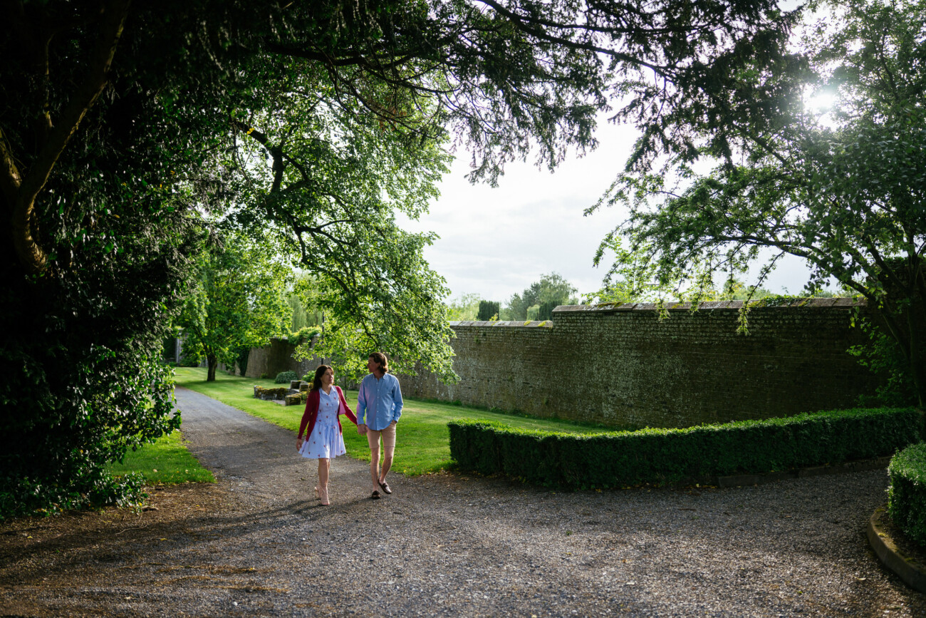 A couple walks hand in hand along a gravel path in a lush garden at Kilkea Castle, surrounded by trees, hedges, and a stone wall—a charming scene perfect for an engagement or proposal.