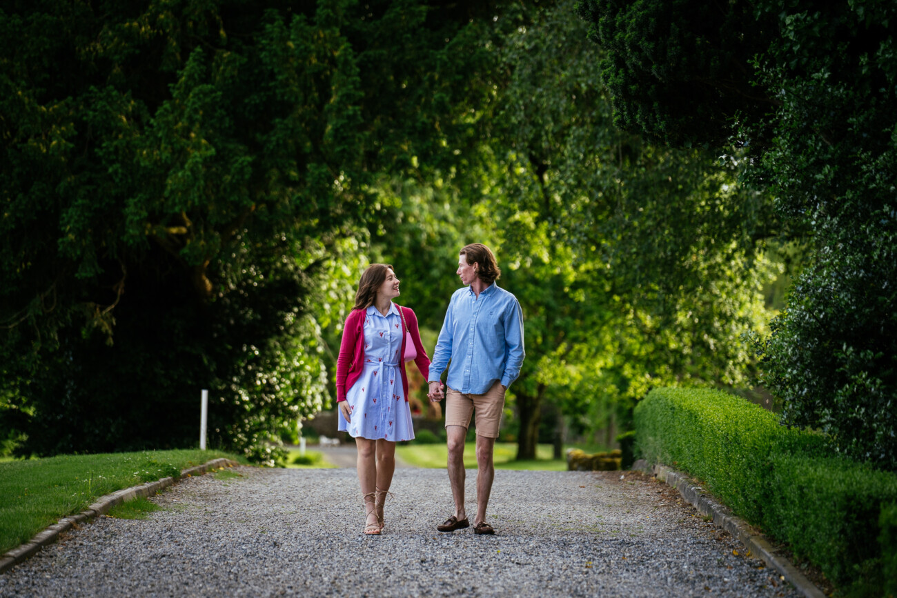 A couple walks hand in hand down a gravel path surrounded by lush green trees and bushes on a sunny day, celebrating their engagement.