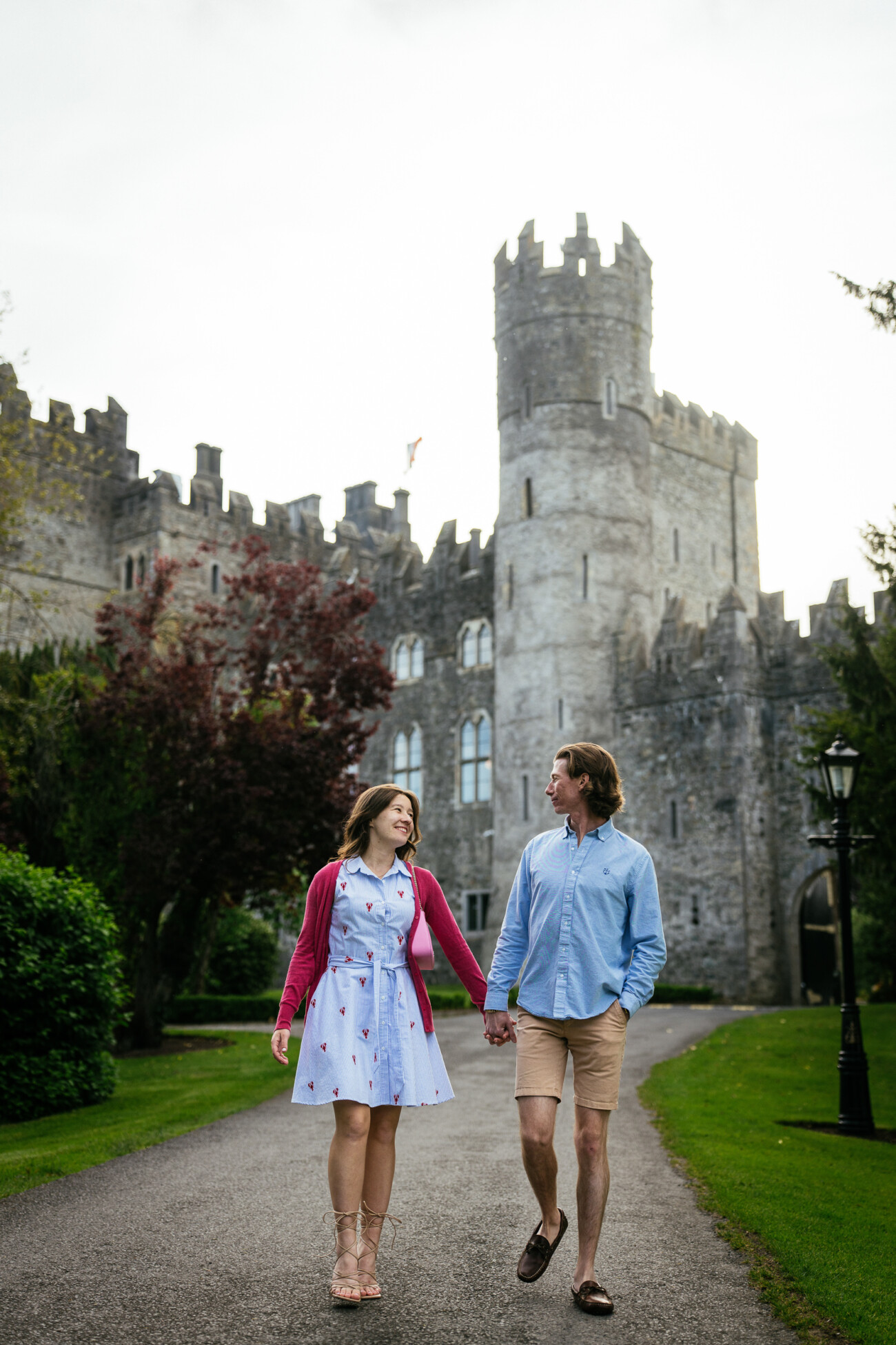 A man and woman hold hands while walking on a path in front of Kilkea Castle, surrounded by greenery—a romantic setting perfect for an engagement or proposal.