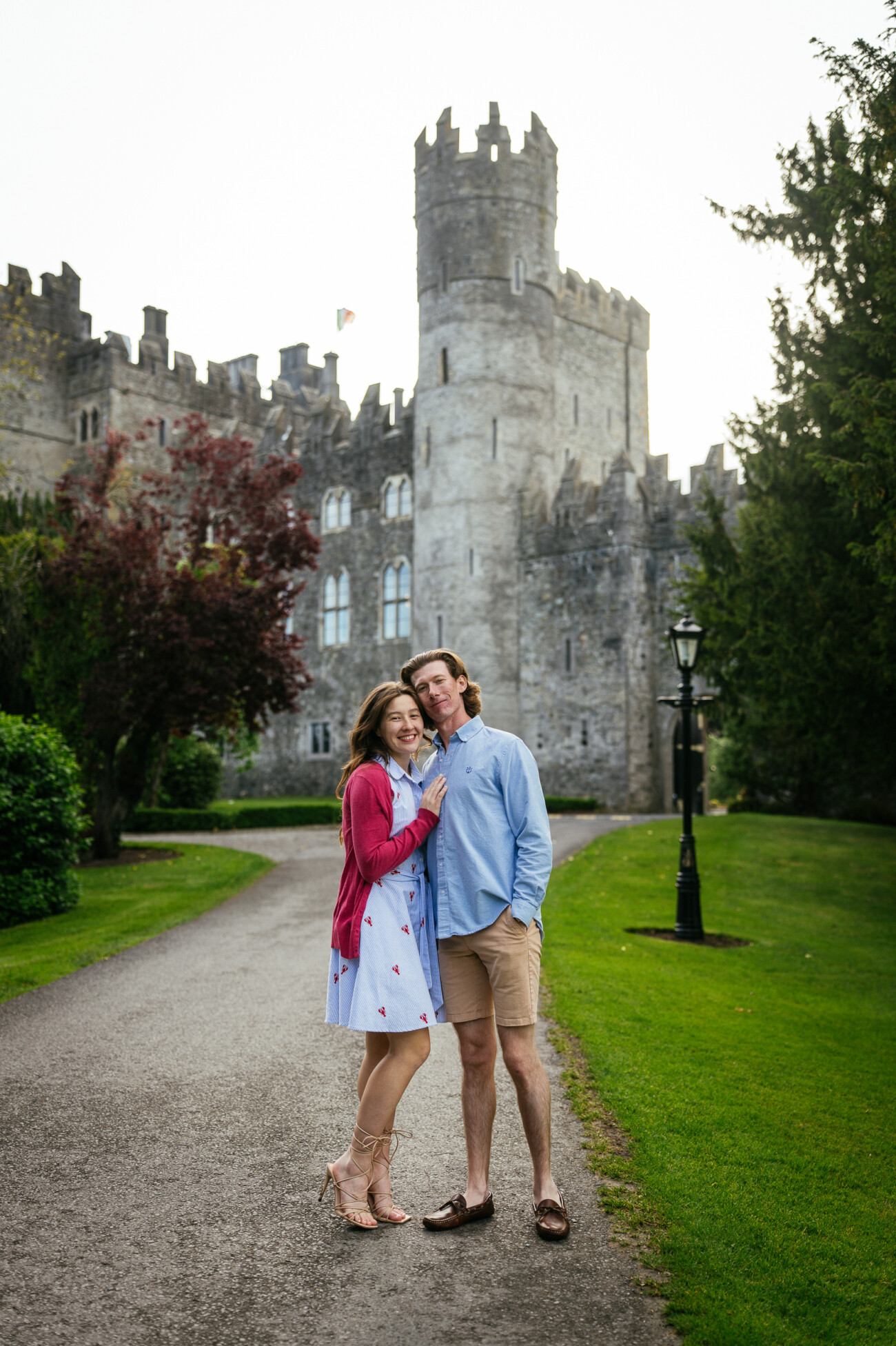 A couple stands on a path in front of Kilkea Castle, surrounded by greenery and trees on a cloudy day—an enchanting setting for an engagement.