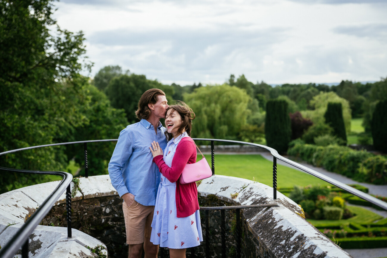 A man and woman stand on a stone balcony at Kilkea Castle overlooking a landscaped garden. The man kisses the woman's head while she smiles, both dressed in casual summer clothing, celebrating their engagement.
