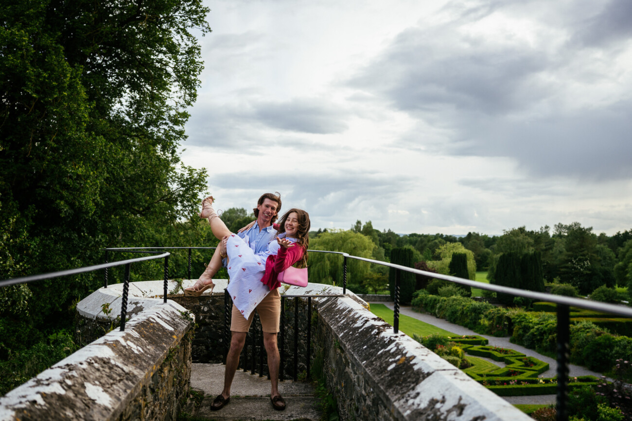 A man stands on a stone walkway at Kilkea Castle, holding a smiling woman in his arms after a magical proposal, with gardens and greenery in the background under a cloudy sky.