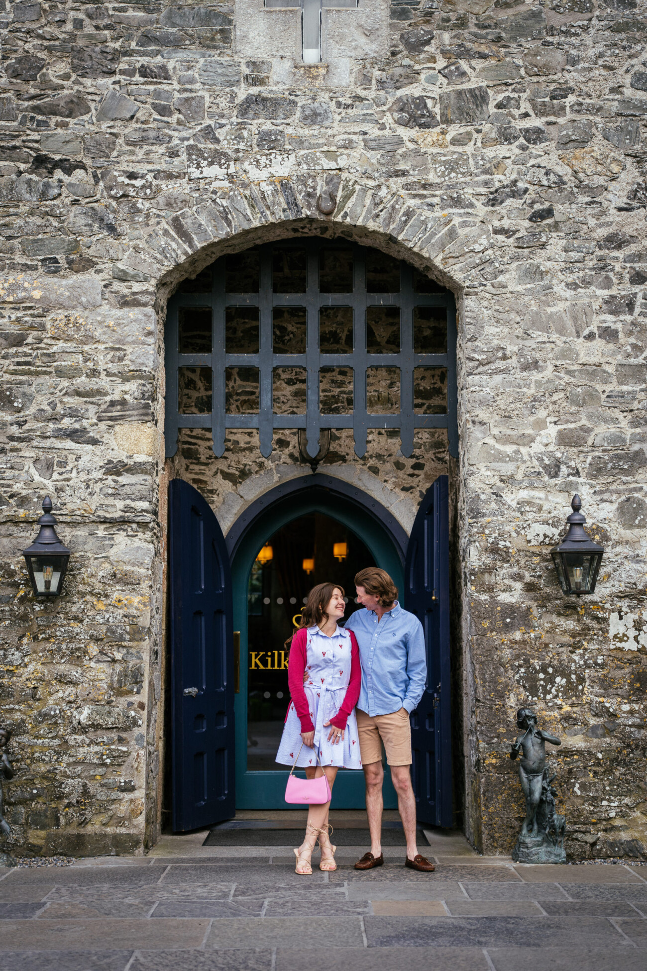 A woman and a man stand together in front of a large stone archway with dark blue double doors and lanterns on each side, capturing the magic of their engagement at Kilkea Castle.