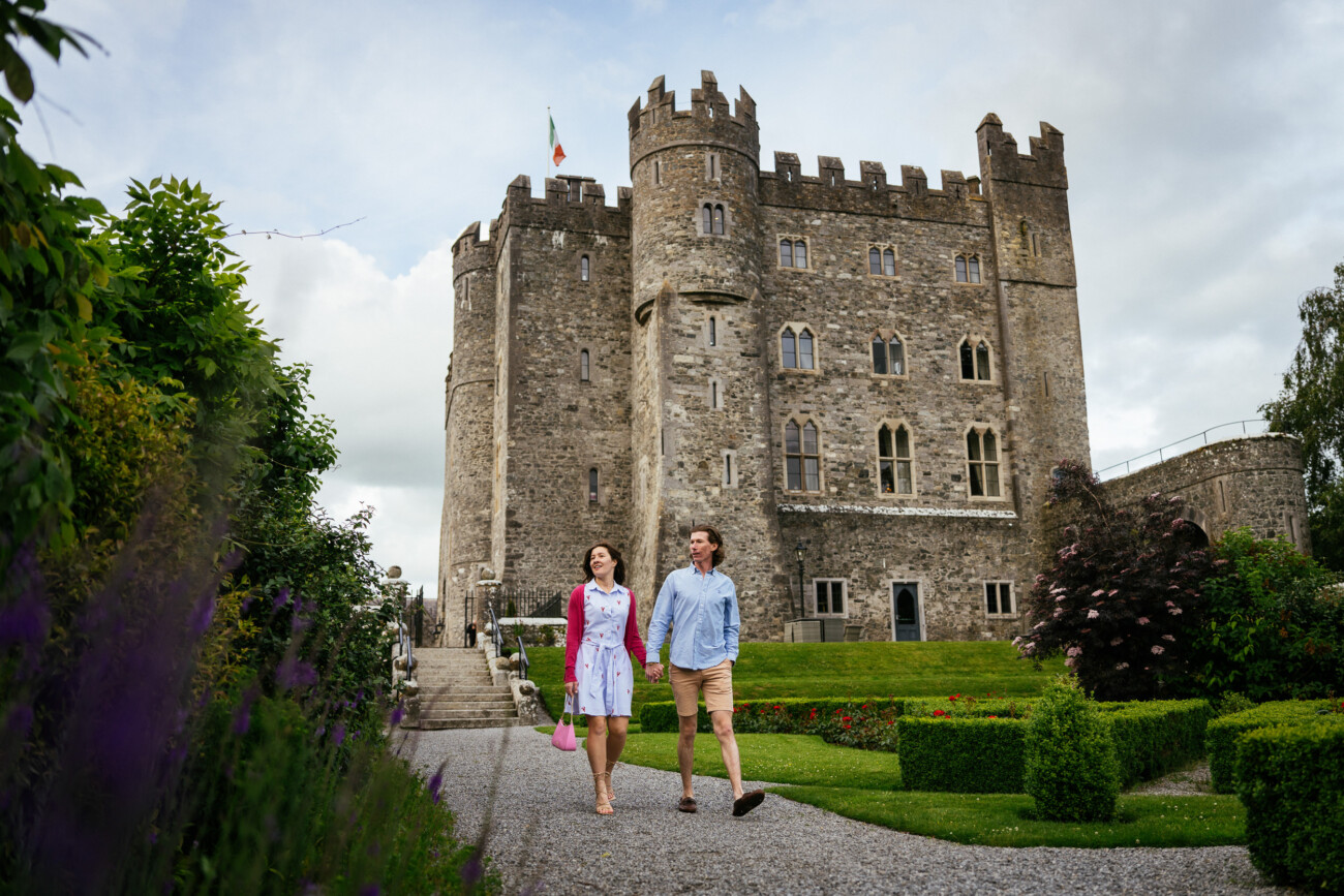 A couple walks along a garden path in front of Kilkea Castle with a flag on top, surrounded by greenery and manicured bushes, the perfect setting for a romantic proposal or engagement.