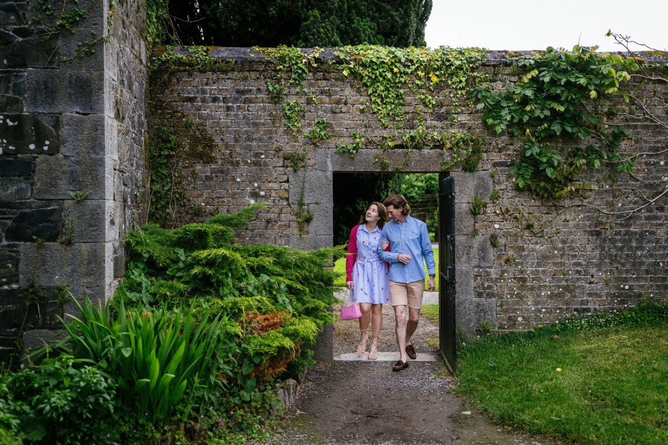 A couple celebrating their engagement walks arm-in-arm through a stone archway surrounded by green plants and ivy-covered walls in the Kilkea Castle garden.