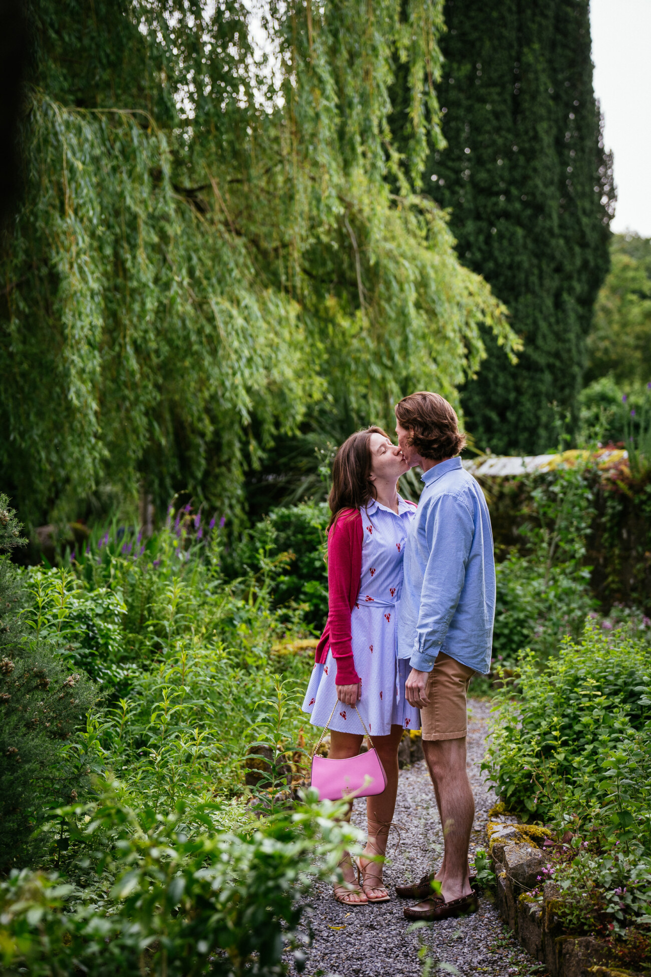A couple stands on a gravel path surrounded by green plants and trees at Kilkea Castle, sharing an engagement kiss. The woman holds a pink bag and wears a light dress with a red cardigan; the man is in a blue shirt and shorts.