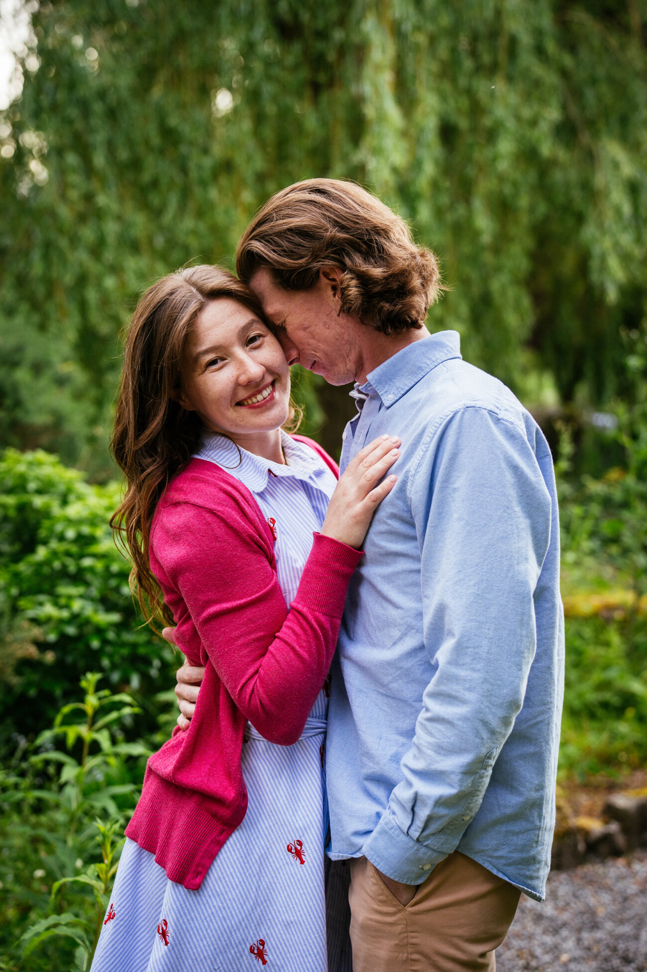 A woman in a red jumper and a man in a light blue shirt embrace outdoors, surrounded by greenery and trees at Kilkea Castle. The woman is smiling at the camera, capturing a joyful engagement moment.