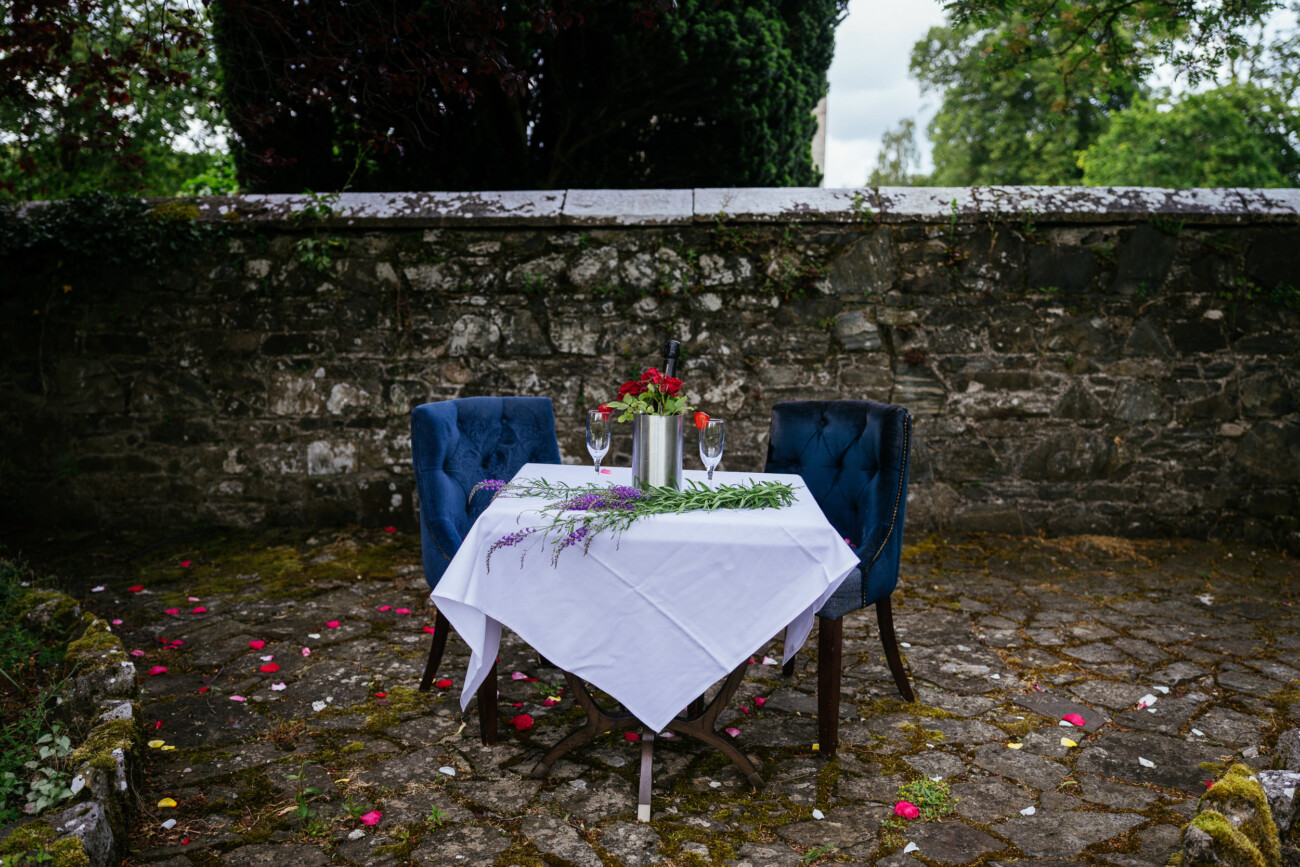 A small outdoor table with a white tablecloth, two blue chairs, flowers, and wine glasses is set on a stone patio near a stone wall—perfect for an engagement or proposal inspired by the romance of Kilkea Castle, with petals scattered on the ground.