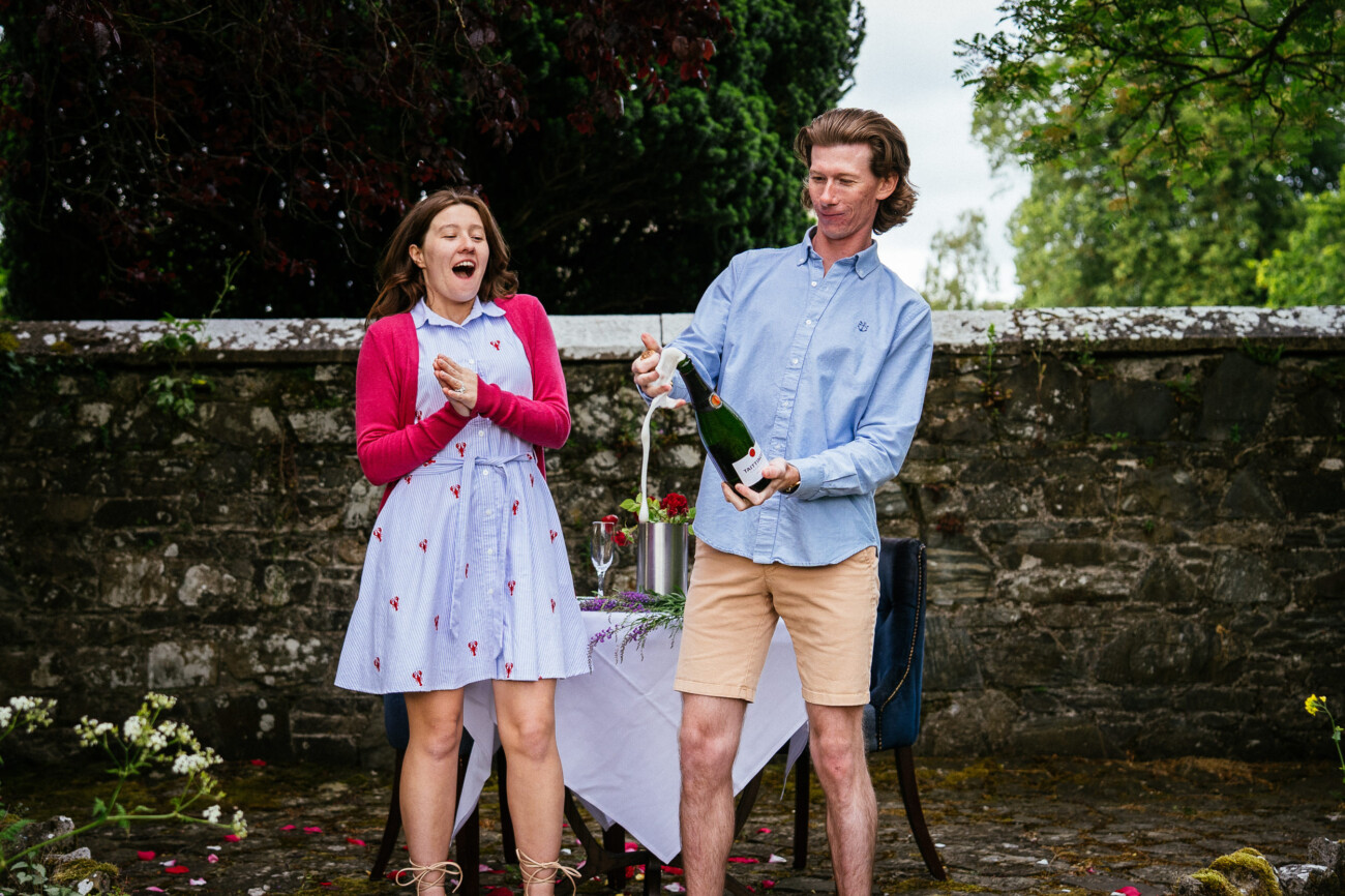 A man opens a bottle of sparkling wine, causing it to froth over, while a woman beside him reacts with surprise—capturing the excitement of an engagement near a stone wall and outdoor table at Kilkea Castle.