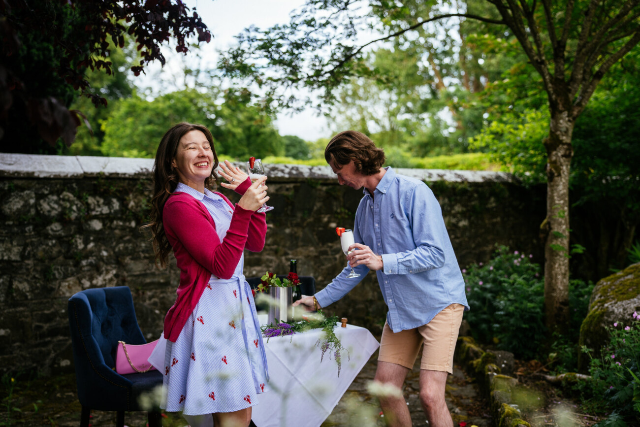 A woman in a blue dress and red cardigan laughs and holds a bottle, whilst a man in a blue shirt arranges a candle on an outdoor table set for their engagement celebration.