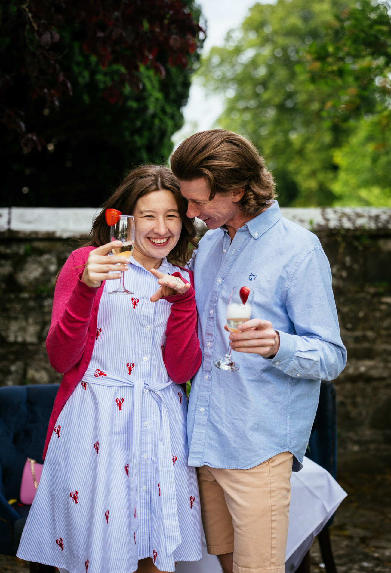 A woman and man stand close together outdoors at Kilkea Castle, holding drinks and smiling. The woman gestures playfully whilst the man looks at her, celebrating their engagement amid greenery, a table, and a stone wall in the background.