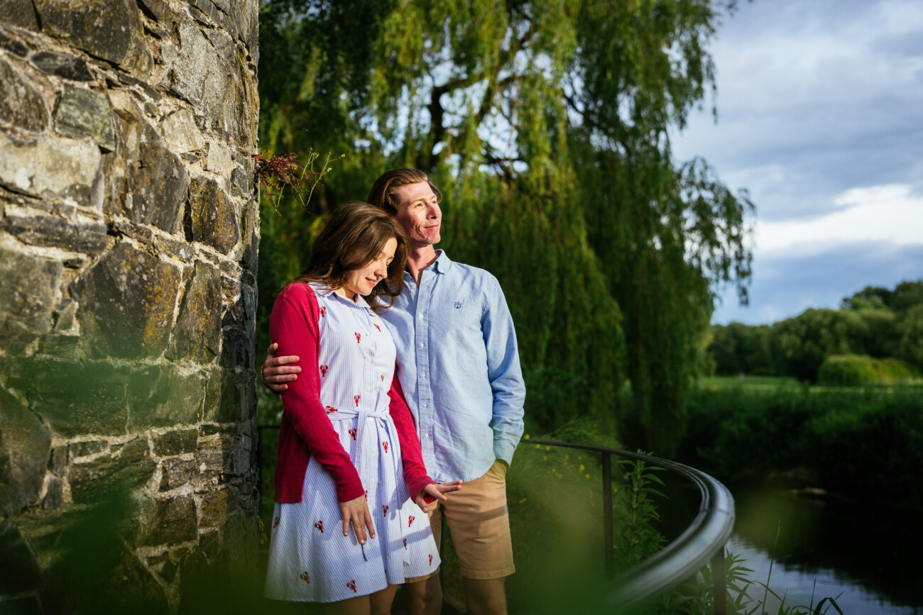 A couple stands by a stone wall and black railing at Kilkea Castle, surrounded by greenery. The woman looks down whilst the man looks ahead, both dressed in casual summer clothes, capturing a quiet proposal moment.