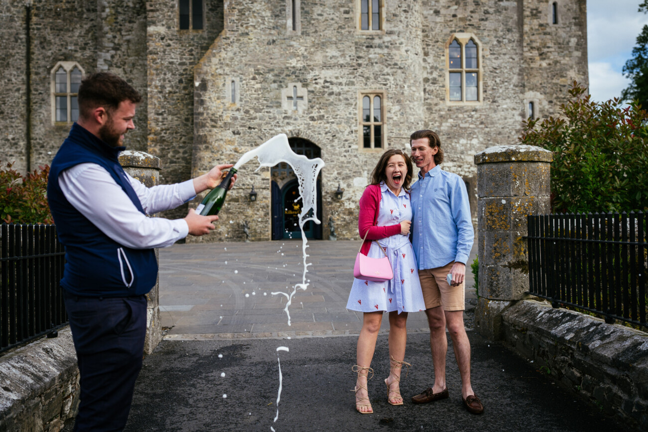 A man pops a champagne bottle, spraying foam, during an engagement photoshoot as a surprised woman and a smiling man pose in front of the historic Kilkea Castle.