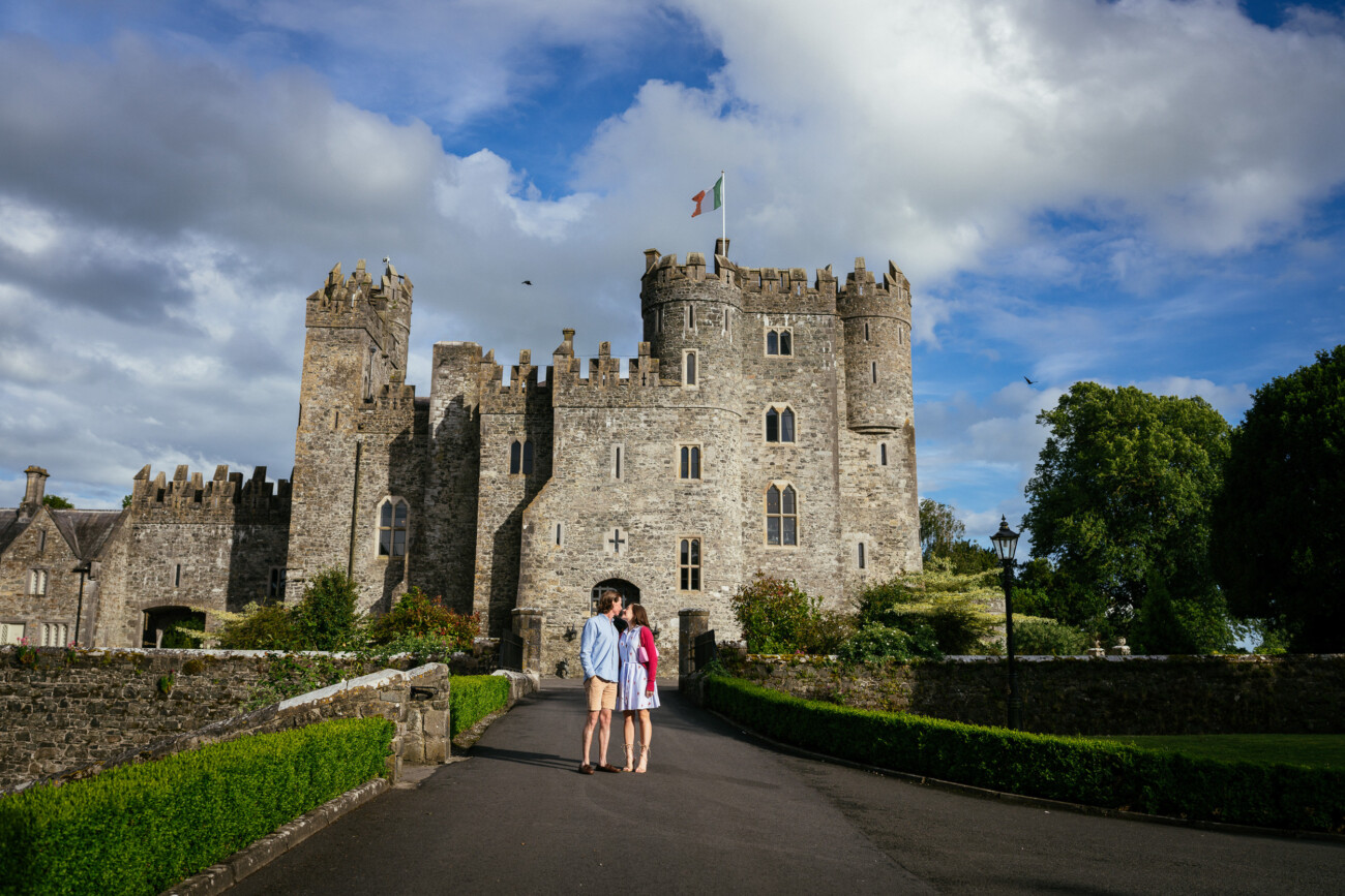 Three people stand in front of Kilkea Castle, its flag flying high, surrounded by gardens and a partly cloudy sky—the perfect setting for an unforgettable engagement photoshoot.