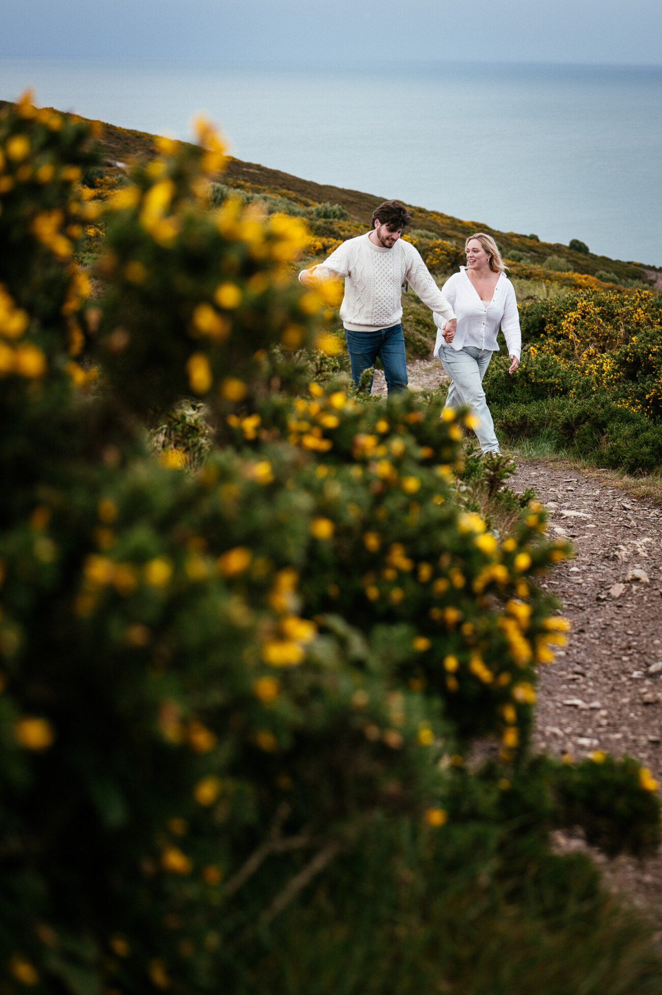 A man and a woman walk hand in hand along a rocky path surrounded by greenery and yellow flowers, with the sea visible in the background.