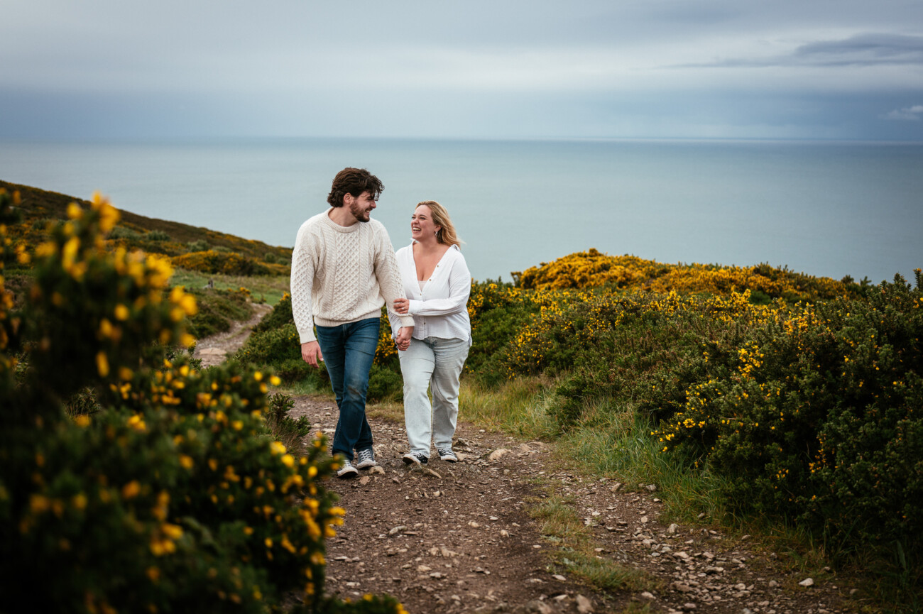 A couple walk together on a rocky path surrounded by green bushes with yellow flowers, overlooking the sea under a cloudy sky.