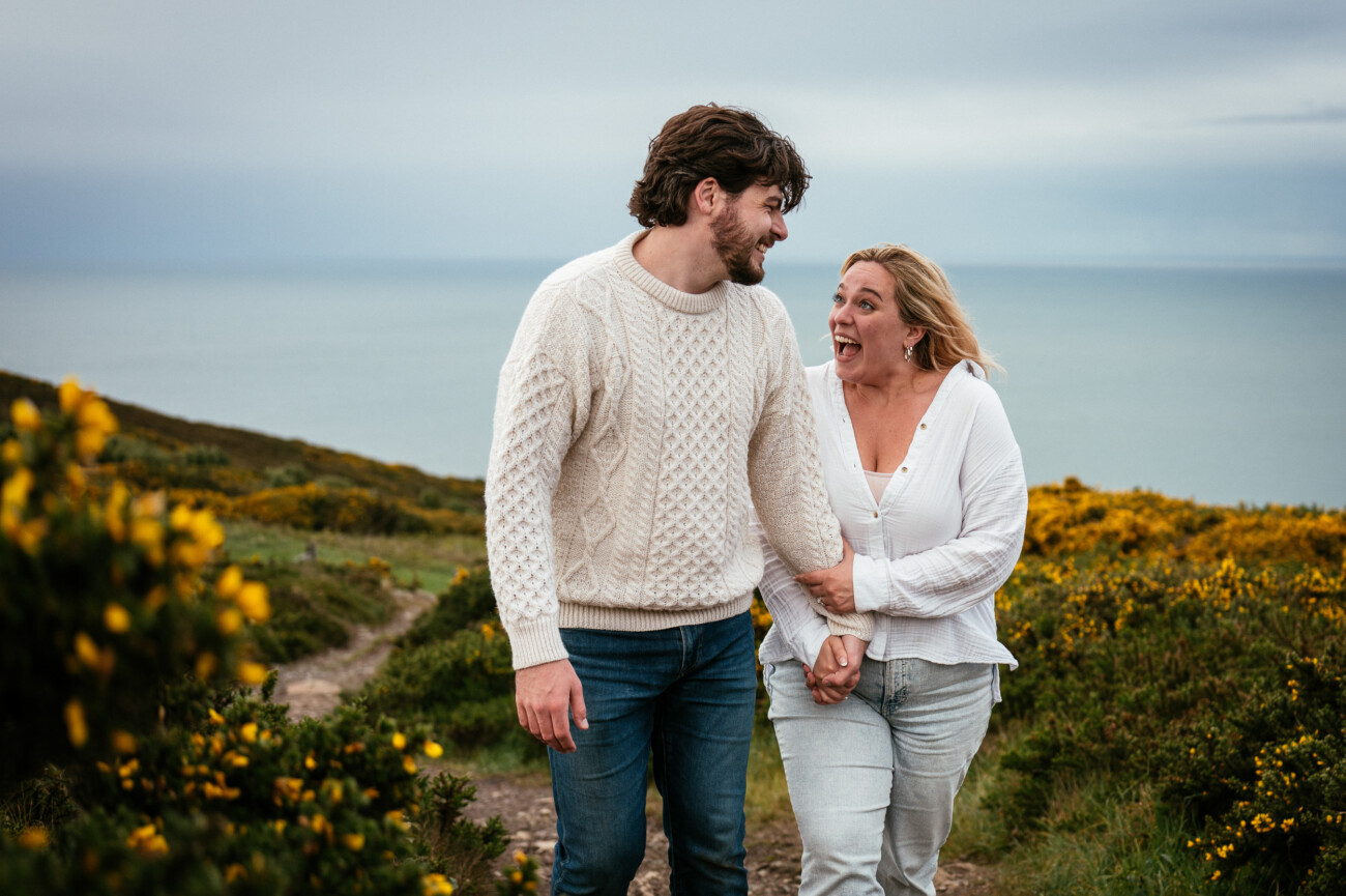 A man and woman walk hand-in-hand along a coastal path with the sea in the background, both smiling and wearing light jumpers and jeans.