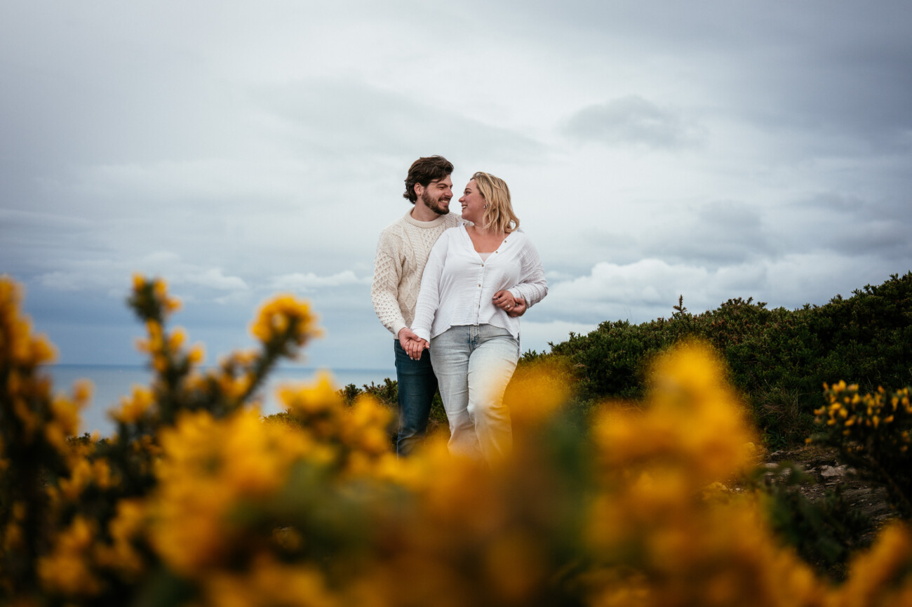 A man and woman holding hands and walking outdoors, surrounded by yellow flowers with a cloudy sky in the background following their secret proposal howth cliffs dublin.