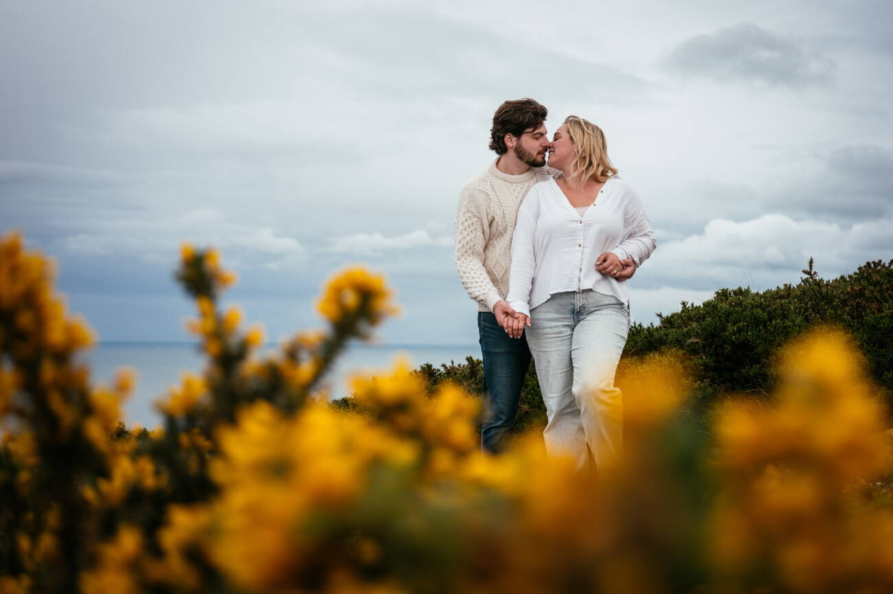 A couple walks hand-in-hand outdoors, surrounded by yellow flowers and greenery under a cloudy sky.