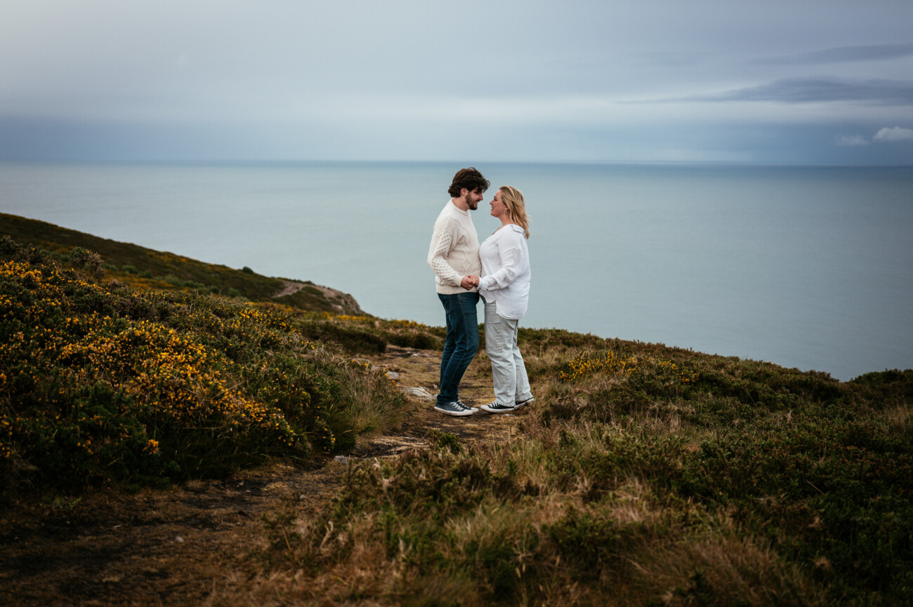 A man and woman stand facing each other on a grassy coastal cliff, holding hands, with the sea and cloudy sky in the background.