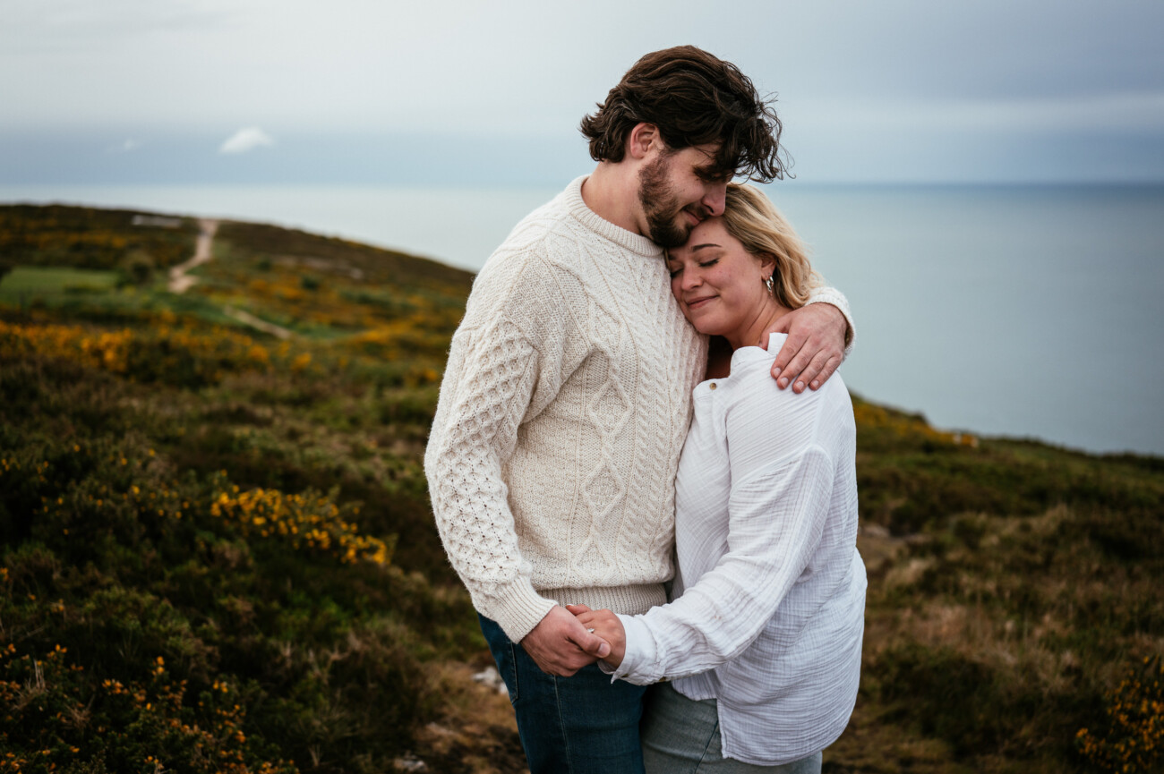 A couple stands embracing and smiling with eyes closed on a grassy hillside overlooking the sea under a cloudy sky.
