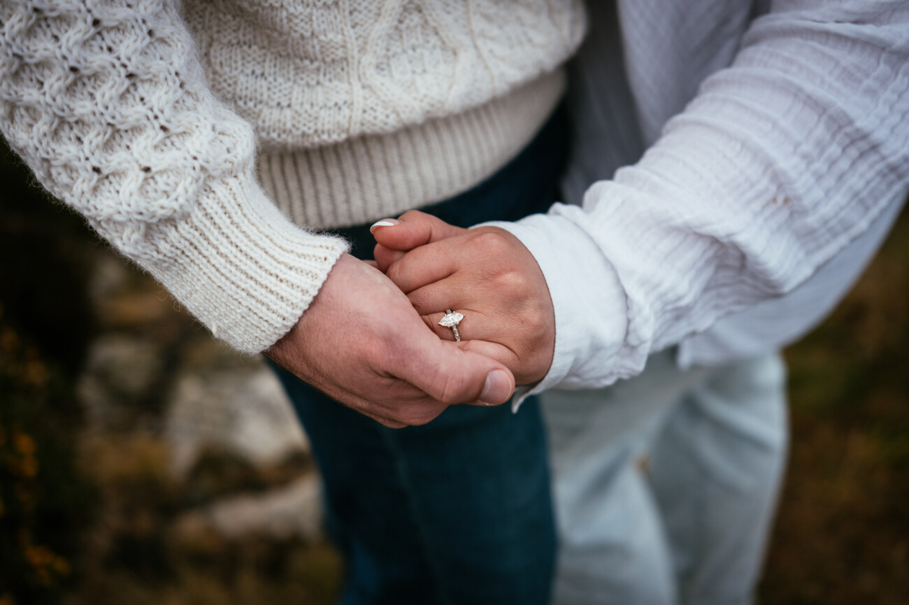 Two people holding hands, one wearing a white jumper and jeans, the other in a white top with a diamond engagement ring on their finger.