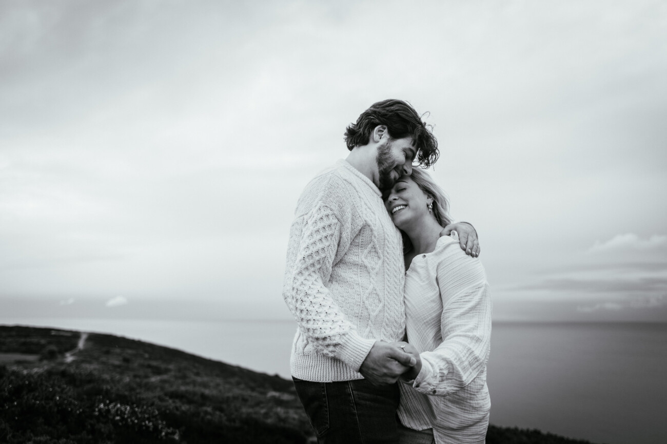 A couple stands on a grassy hill near the sea, embracing and smiling at each other, with cloudy skies in the background.