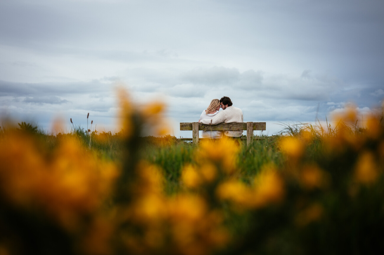 Two people sit closely together on a bench in a grassy field, facing away from the camera, with yellow flowers blurred in the foreground and a cloudy sky above.