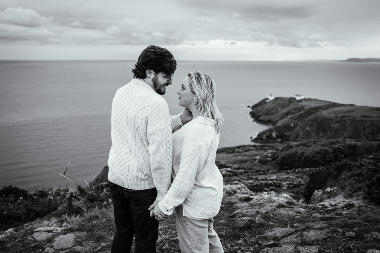 A couple stands on a rocky cliff overlooking the sea, holding hands and facing each other, with a lighthouse visible in the distance.
