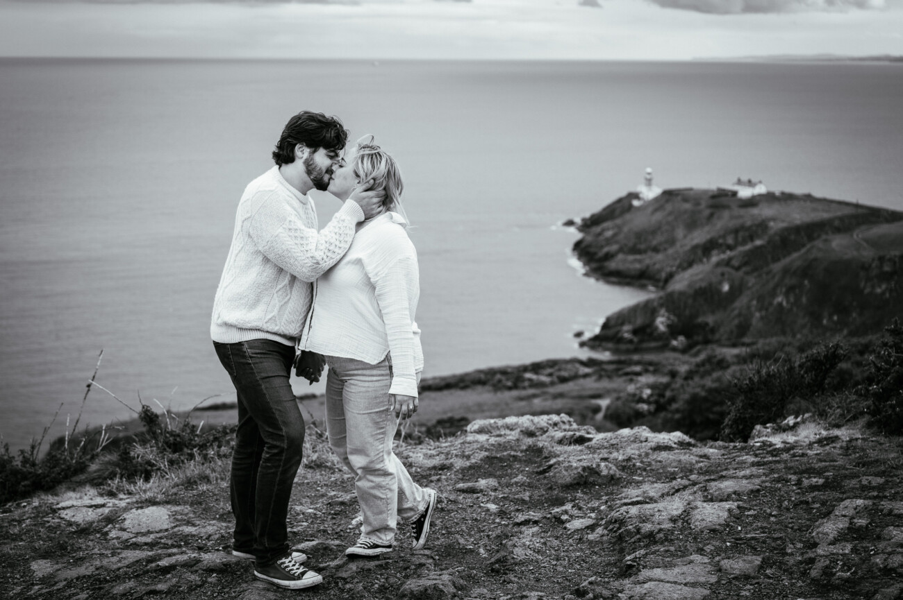 A man kisses a woman on the forehead whilst standing on a rocky cliff overlooking the sea, with a lighthouse visible in the background. The photo is in black and white.