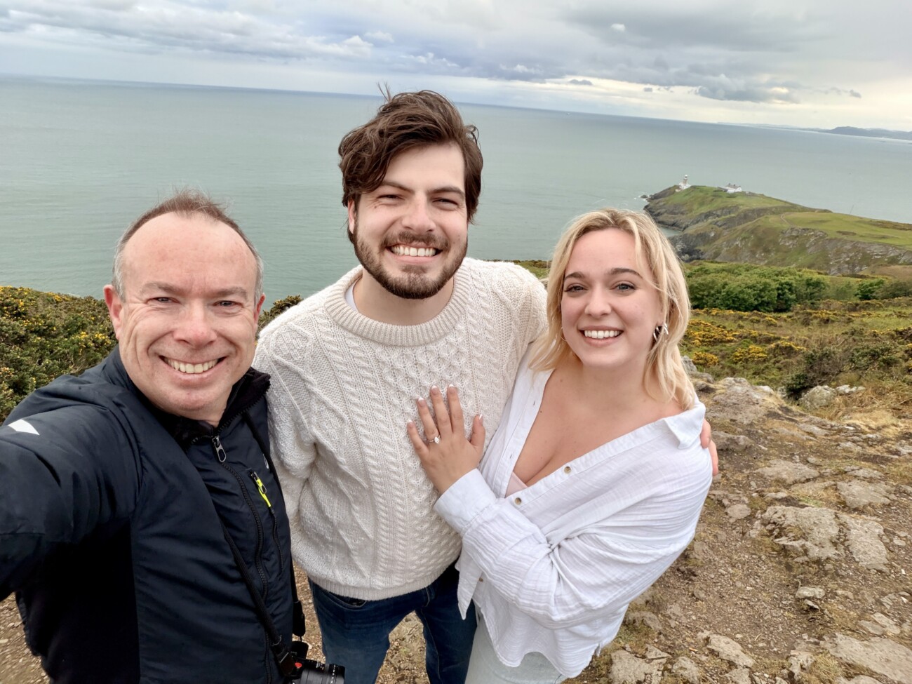 Three people stand smiling on a rocky cliff with the sea and coastline in the background, under a cloudy sky.