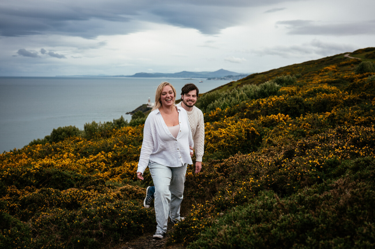 Two people walk up a hillside covered in green and yellow vegetation with the sea and distant land visible under a cloudy sky.