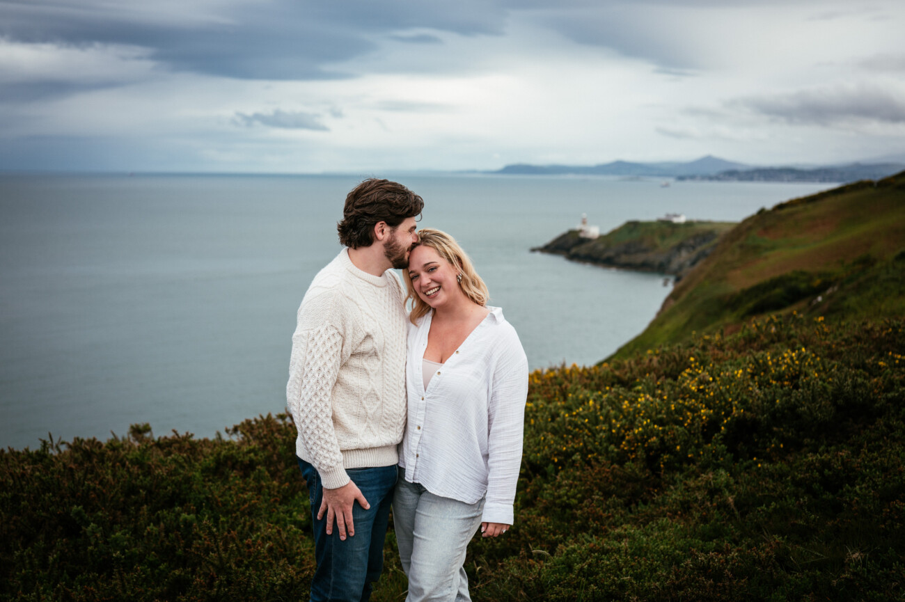 A couple stands on a grassy clifftop overlooking the sea, with the man kissing the woman's forehead. Cloudy sky and distant coastline are visible in the background.