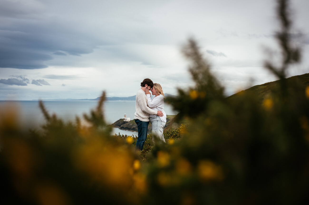 A couple embraces and touches foreheads on a grassy hilltop, with the sea and a distant lighthouse in the background and blurred yellow flowers in the foreground following their secret proposal howth cliffs dublin.