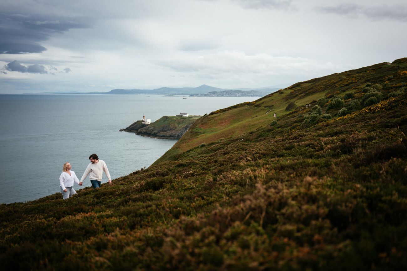 Two people walk up a grassy hillside overlooking the sea, with a lighthouse visible in the distance under a cloudy sky.