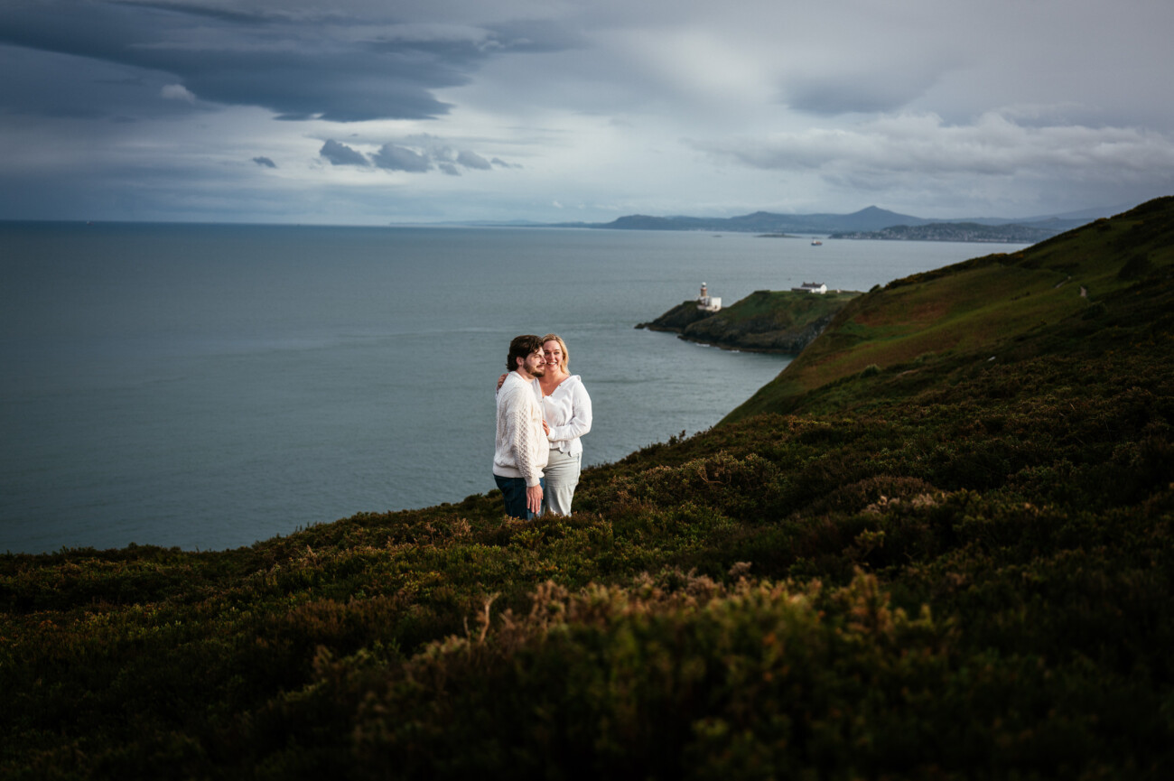 Two people stand close together on a grassy hillside overlooking the Irish sea, with Bailey lighthouse on a distant headland and mountains in the background under a cloudy sky following their secret proposal howth cliffs dublin.