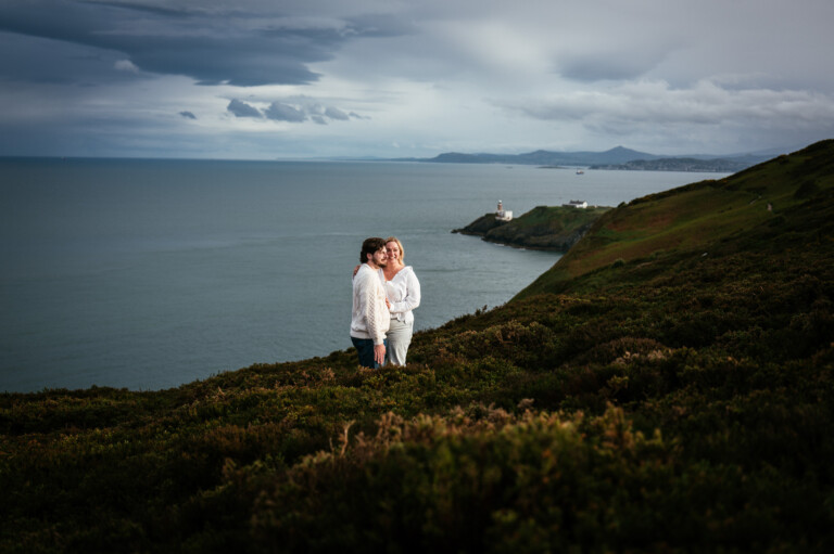 Two people stand close together on a grassy hillside overlooking the Irish sea, with Bailey lighthouse on a distant headland and mountains in the background under a cloudy sky following their secret proposal howth cliffs dublin.